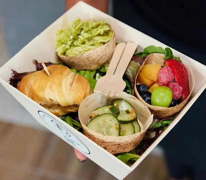 A Knife & Faulk boxed lunch with a croissant sandwich, guacamole, fresh fruit and cucumber salad